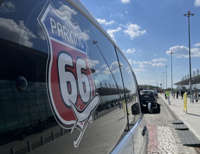 Parkplatz 66 - Ein glänzendes Auto mit Logo auf dem Parkplatz. Sonniger Tag mit bewölktem Himmel. Im Hintergrund sind Gebäude und Straßenlaternen sichtbar.