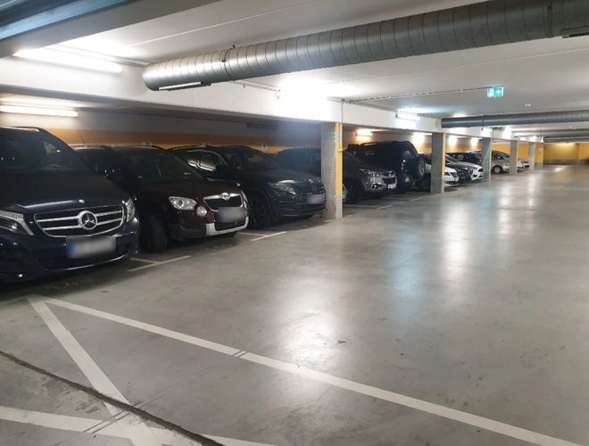 Podziemny parking z zaparkowanymi samochodami wzdłuż ściany, dobrze oświetlony przestronny korytarz.