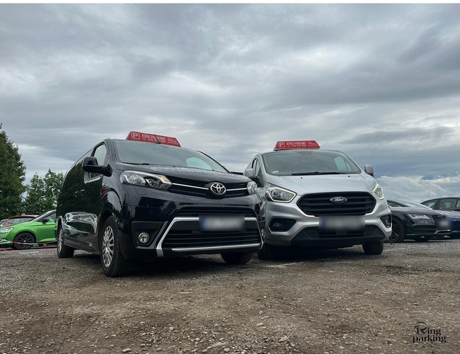 Parkovisko v Krakove KING - Dve veľké autá stoja vedľa seba. Pozemok je štrkový a otvorený. Tmavé mraky na oblohe.