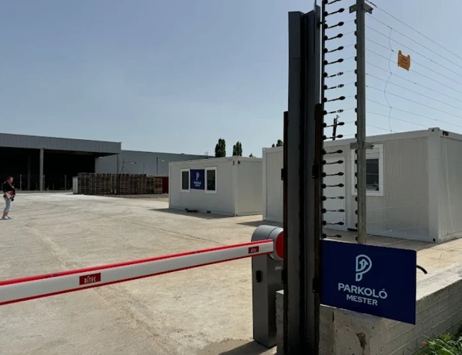 Szlaban wjazdowy na teren parkingu widziany z bliska, obok znajduje się ogrodzenie oraz biały kontener biurowy ustawiony na utwardzonym placu