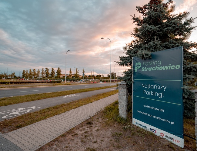 Parking Strachowice – obok stoi oznakowany baner. W tle biegnie droga i ścieżka rowerowa. Rośnie rząd drzew.