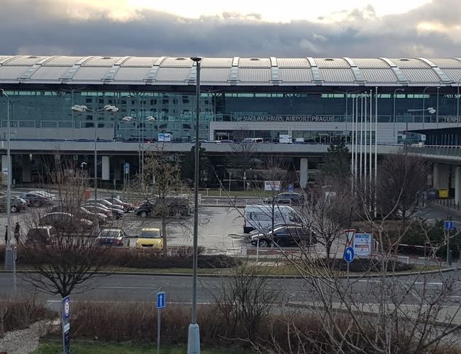 Parking Smile Parking – widoczny jest duży, otwarty parking. W tle znajduje się nowoczesny budynek terminalu lotniska. Atmosfera jest pochmurna.