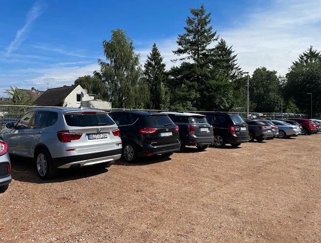 Parking Park In Fly Out - Kilka samochodów stoi na żwirowym placu. Teren jest ogrodzony. W tle widoczne są drzewa i budynek.