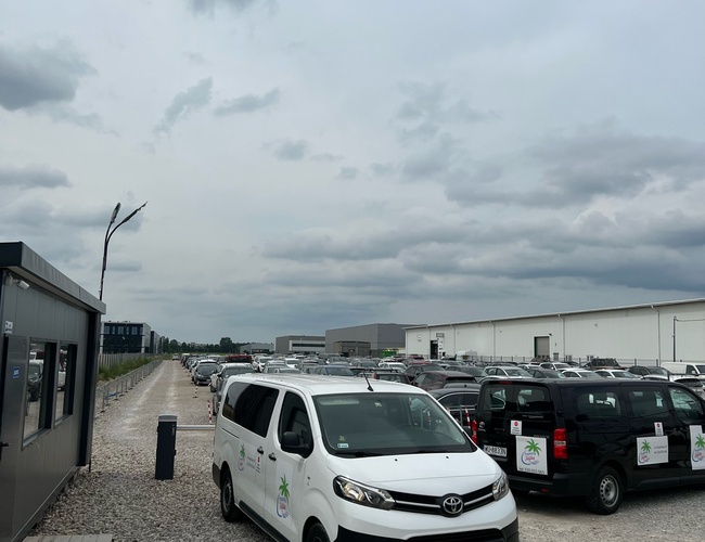 Holiday Parking – Parking jest nieogrodzony i żwirowy. Widać zaparkowane samochody i kontener biurowy.