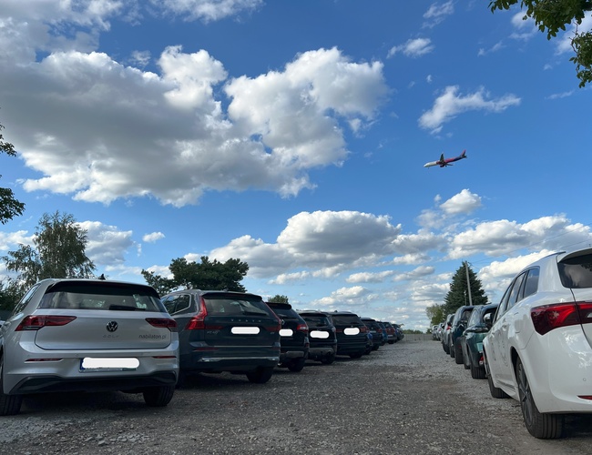 PRO-PARK – Samochody stoją równo na żwirowym parkingu. Nad nimi leci samolot. Pogodne niebo z chmurami.