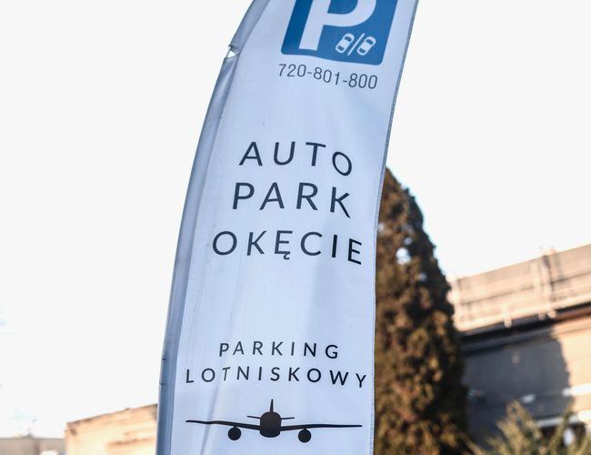 Auto Park Okęcie – Flaga reklamowa przy wejściu na parking. W tle widoczny jest czarny samochód i budynek.