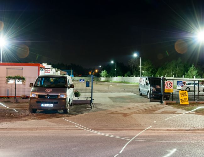 AAP-66 – Parking ogrodzony, dobrze oświetlony. Wjazd z bramą. Dwa zaparkowane pojazdy, wolne miejsca dostępne.