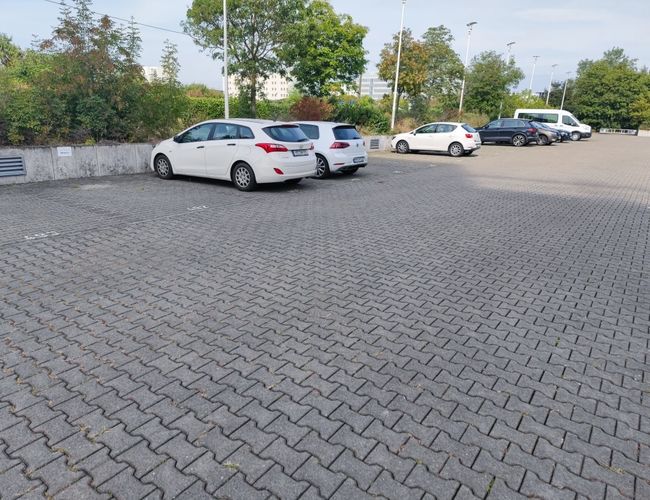 Parking FrankfurtValet24 – Kilka samochodów stoi na utwardzonej powierzchni. W tle roślinność i latarnie. Plac jest częściowo zapełniony.