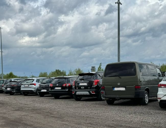 Parking 66 - Szereg samochodów stoi na żwirowej nawierzchni. Latarnie oświetlają teren. W tle widać drzewa i ogrodzenie.