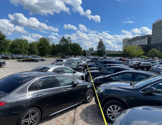 Parking Smile - Na zdjęciu widoczne są auta na żwirowym placu. Parking jest otwarty i przestronny. W tle widać drzewa i budynki.