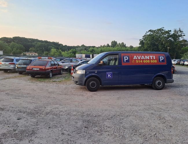 Estacionamiento Avanti - En el área de grava hay varios autos estacionados. Al fondo se pueden ver árboles y mucha vegetación. Junto a los autos hay una furgoneta azul con el logo del estacionamiento.
