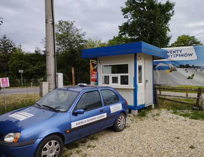 Parking Guardia - Niebieski samochód stoi przy budce ochrony. Budka ma białe ściany i niebieski dach. W tle jest reklama nad piaskowym placem.
