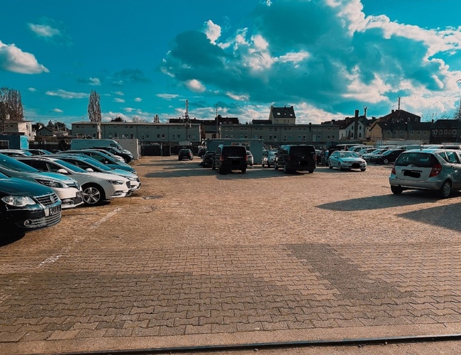 Parkovacie bezpečnostparking24 VALET – Veľké, otvorené parkovisko. Automobily sú zoradené v radoch. V pozadí sú viditeľné budovy.