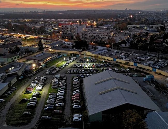 Parqueo en el Aeropuerto Parktiger 2 - vista de un gran estacionamiento iluminado. Los coches están alineados en filas ordenadas. En el fondo se ven los edificios de la ciudad.