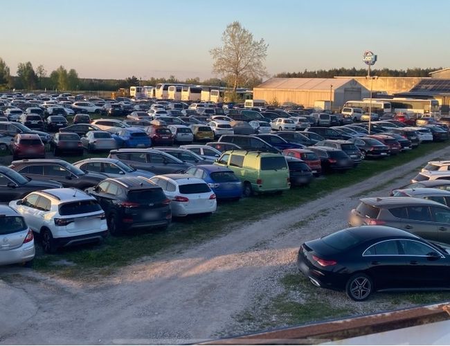 Aparcamiento bajo el molino – Muchos coches aparcados en una superficie pavimentada. Al fondo se pueden ver árboles y edificios.