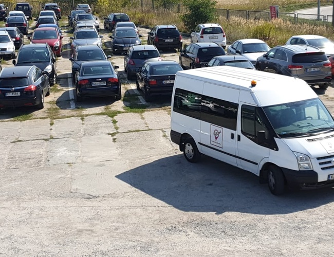 JG Parking – Varios filas de coches aparcados. La parte delantera de una furgoneta blanca en primer plano. Área cubierta de losas de hormigón.