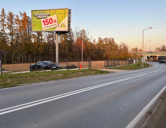 Victory Cars - Parking ogrodzony siatką z bramą. Przy drodze stoją latarnie. Obok widać reklamę na bilbordzie.