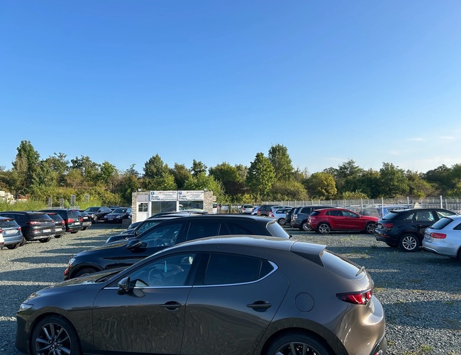 Estacionamiento de chófer de lanzadera – Hay coches en el estacionamiento. El área está cercada. Al fondo hay árboles.