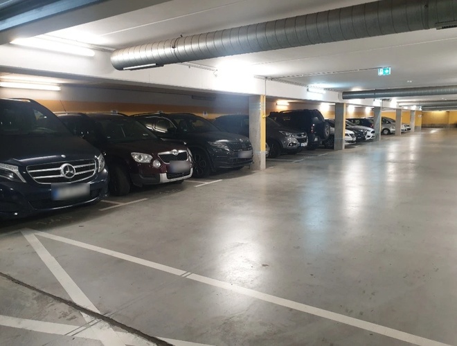 Podziemny parking z zaparkowanymi samochodami wzdłuż ściany, dobrze oświetlony przestronny korytarz.