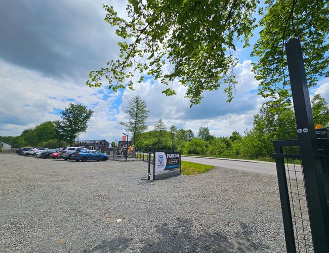 ParkZone – na zdjęciu jest żwirowy parking z kilkoma samochodami. Teren jest ogrodzony i otoczony drzewami. Pogoda jest pochmurna z przejaśnieniami.