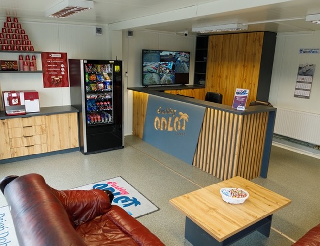 Parkovisko Odlet – recepcia s pohovkou a automatom na nápoje. Na pulte ležia letáky. V pozadí je vidieť monitor.