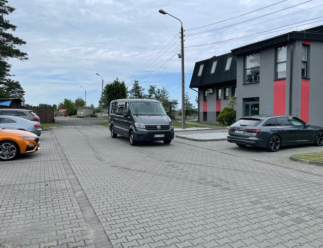 Parking Port - Na placu stoją zaparkowane trzy auta. Obok znajduje się nowoczesny budynek z oknami. W tle widać drzewa i niebo.