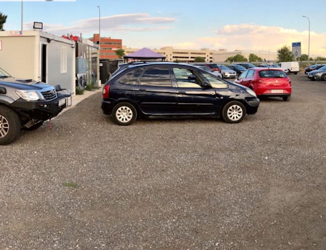 Parking Elite Valet - En el área de grava hay muchos coches. Al lado hay un contenedor de oficina blanco. Al fondo hay árboles y edificios.