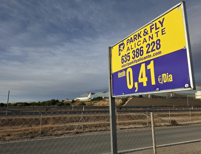 Tablica reklamowa parkingu Park & Fly przy drodze, z informacją o niskiej cenie parkowania oraz numerem telefonu, na tle otwartego terenu i nieba.