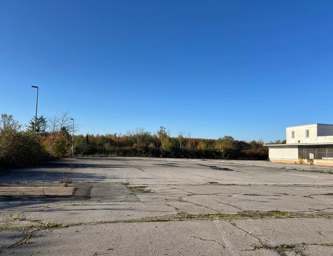 Parking My Travel Carpark Valet – Pusty teren z pękniętym asfaltem. Jeden budynek stoi z boku. W tle widać drzewa i krzewy.