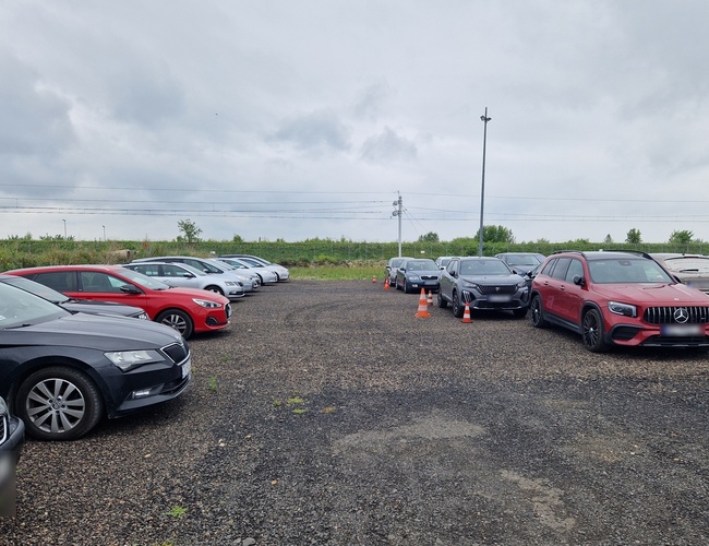 Parking Prime - Coches estacionados en un área de grava. Junto a ellos hay postes de tránsito. Al fondo se ve vegetación y matorrales.