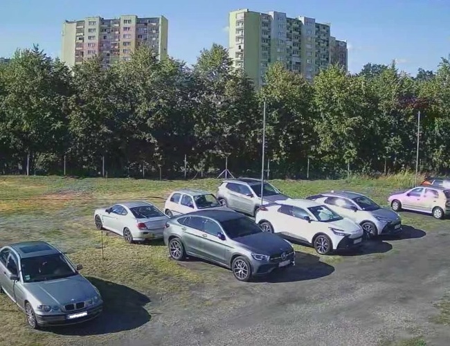 Odlotowy Parking BZG – Na placu stoi kilka aut. Teren jest nieogrodzony i otoczony drzewami. W tle widoczne są bloki mieszkalne.