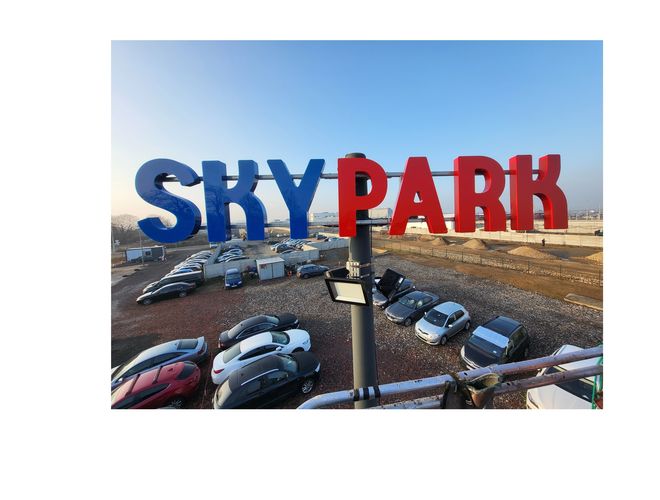 Sky Park – widoczny parking z wieloma samochodami. Teren otoczony jest ogrodzeniem. Neony z napisem są na wysokości.