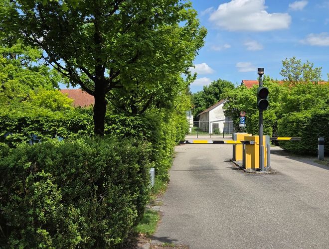 Atomis Hotel München Repülőtéri Parkoló – faösvény az autómatikus sorompó blokkolja a bevezető utat a parkolóba. A háttérben tábla és egy fehér épület.
