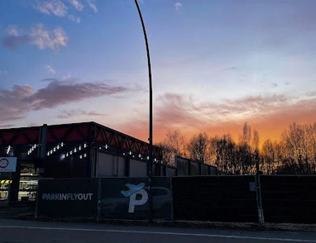 Parking Park In Fly Out – ogrodzony teren z latarnią. W tle widać zachodzące słońce i drzewa. Niebo jest w kolorach zachodu.