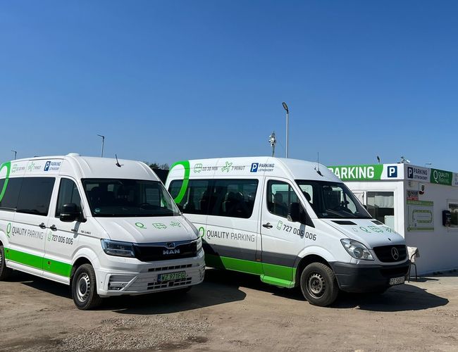 Q-Parking – Dwa białe busy stoją obok budki parkingowej. Parking jest dobrze oświetlony w słoneczny dzień.