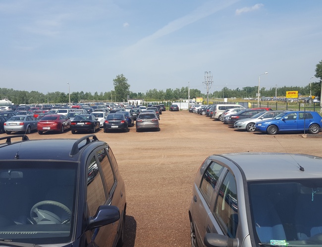 AAP-66 – parking nieogrodzony z licznymi pojazdami. Teren jest piaszczysty, otoczony zielenią. Widoczne są latarnie oświetleniowe.