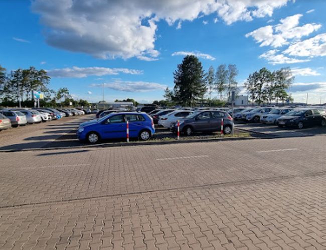 Oficjalny Parking P2 – Na parkingu stoją zaparkowane samochody. Teren jest utwardzony kostką brukową. W tle rosną drzewa.