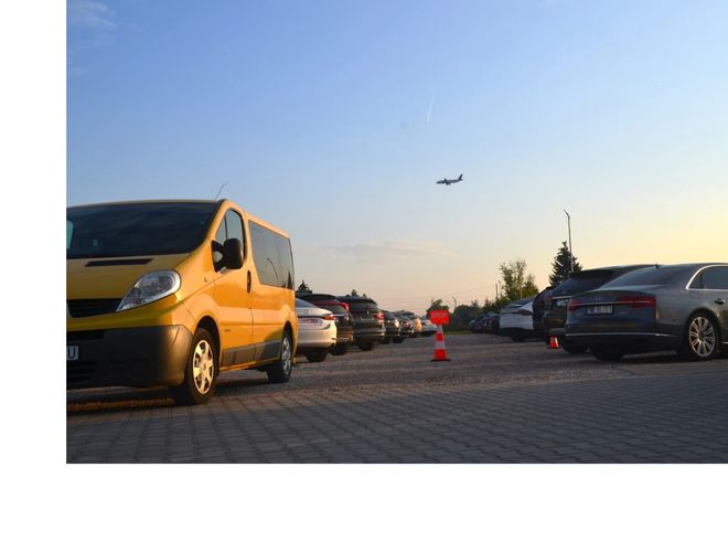 Royal Parking - Na parkingu stoi żółty bus i kilka aut. W tle przelatuje samolot na niebie.