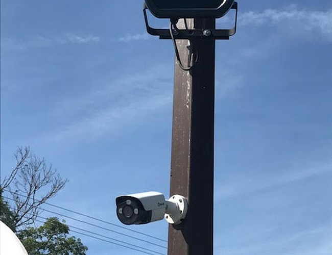 Automat na letisko – Na stĺpe je kamera a lampa. Na pozadí sú vidieť obloha a stromy.