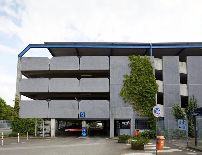 Widok na wielopoziomowy parking od strony frontowej. Widoczna jest betonowa elewacja budynku, główny wjazd na poziomie parteru oraz drzewa i oznakowanie w otoczeniu.