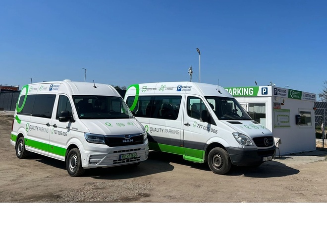 Q-Parking – Dos furgonetas blancas están delante de una cabina con un letrero. El terreno no está pavimentado y hay una cerca de malla visible.