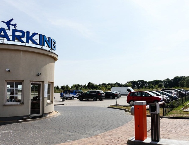 Dostupné a bezpečné parkovisko PREMIUM - na obrázku je vidieť parkovisko s budovou a autami. Vedľa stojí značka s nápisom „Šťastnú cestu!“. Parkovisko je oplotené.