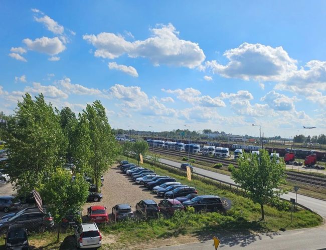VIP Parking - przy trasie S2, teren jest ogrodzony. Widać zaparkowane samochody i rzędy drzew. Na horyzoncie znajdują się tory kolejowe.