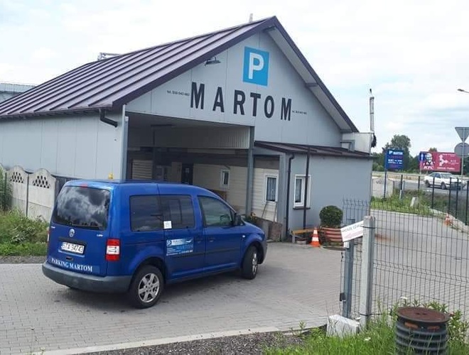 Estacionamiento MARTOM – Un coche azul está frente a una puerta gris. El área está cercada. En el fondo se ven carteles publicitarios.