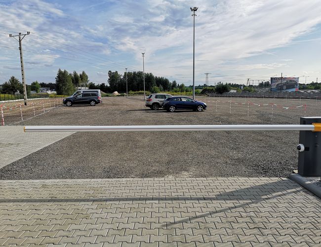 Parking A1 Rondo – duży plac z zaparkowanymi samochodami. Teren jest ogrodzony i żwirowy. Widać barierkę i lampy oświetleniowe.