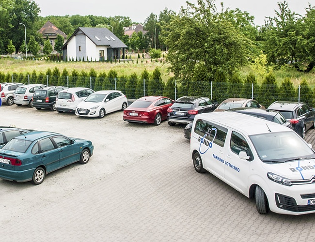 Parking Rondo – Teren jest ogrodzony. Samochody stoją na żwirowej powierzchni. W tle jest dom i zieleń.