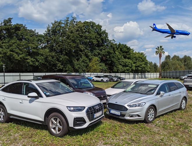AVT60 - Hay varios coches en el aparcamiento. En el fondo, hay un avión volando. El área está rodeada de una cerca y vegetación.
