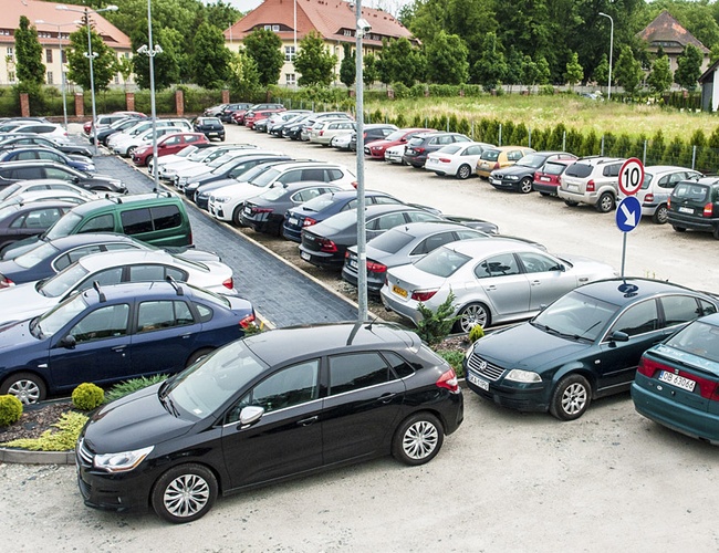 Parking Rondo - Na zdjęciu widać wiele zaparkowanych samochodów. Teren otacza niska roślinność. Naprzeciwko widoczne są budynki.