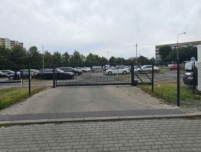 Odlotowy Parking BZG - Widok na zamkniętą, ogrodzoną bramę parkingu. W tle stoją zaparkowane samochody. Wokół rosną drzewa.