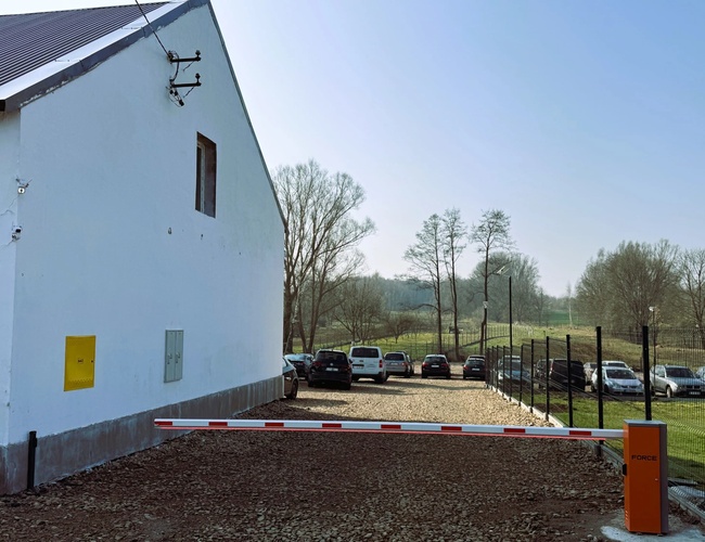 Parkovisko Amber – pred bielou budovou stojí závor. Zaparkované autá sú viditeľné za oplotením. Vedľa parkoviska rastú stromy.